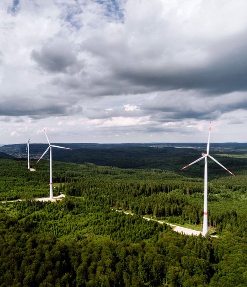 Drei Windkraftanlagen stehen in einem grünen Waldgebiet. Die Anlagen sind hoch, mit weißen Turbinen und roten Akzenten. Der Himmel ist bewölkt, und im Hintergrund sind sanfte Hügel zu sehen. Die Landschaft ist von dichten Bäumen umgeben, die eine satte grüne Farbe haben.