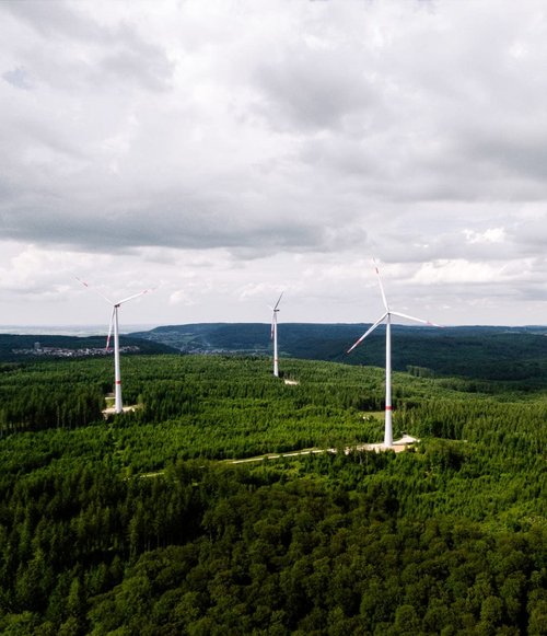 Eine Luftaufnahme von mehreren Windkraftanlagen, die in einem dichten, grünen Wald stehen. Die fünf Anlagen sind auf einer bewaldeten Anhöhe platziert, umgeben von Bäumen. Der Himmel ist bewölkt, und im Hintergrund sind sanfte Hügel und eine Siedlung zu sehen. Die Windräder haben graue Türme mit roten und weißen Elementen und ihre Rotoren sind in Bewegung.