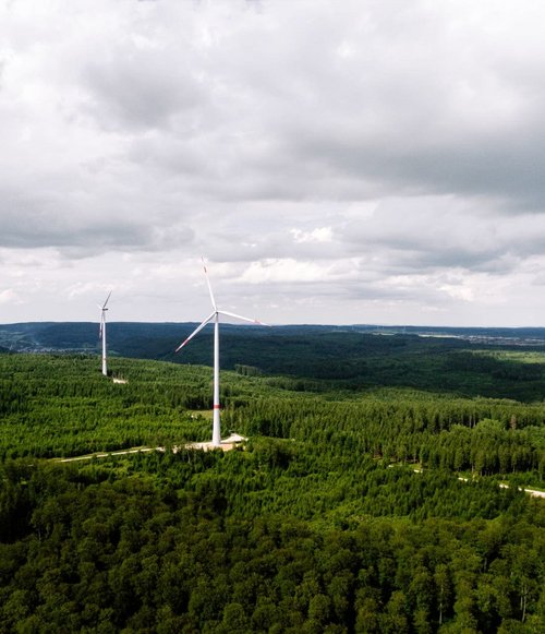 Eine Luftaufnahme von mehreren Windkraftanlagen, die in einem dichten, grünen Wald stehen. Die fünf Anlagen sind auf einer bewaldeten Anhöhe platziert, umgeben von Bäumen. Der Himmel ist bewölkt, und im Hintergrund sind sanfte Hügel und eine Siedlung zu sehen. Die Windräder haben graue Türme mit roten und weißen Elementen und ihre Rotoren sind in Bewegung.