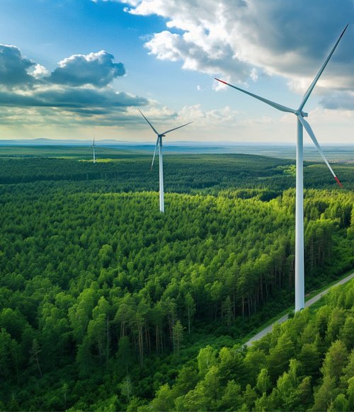 Eine grüne, bewaldete Landschaft mit mehreren Windkraftanlagen, die sich über die Hügel erstrecken. Der Himmel ist teilweise bewölkt, mit einigen Sonnenstrahlen, die durch die Wolken scheinen. Ein geschwungener, schmaler Weg durchzieht den Wald, umgeben von dichtem grünen Laubwerk. Die Windkraftanlagen sind hoch und stehen zwischen den Bäumen, deren Blätter in verschiedenen Grüntönen leuchten. In der Ferne sind weitere bewaldete Hügel zu sehen.